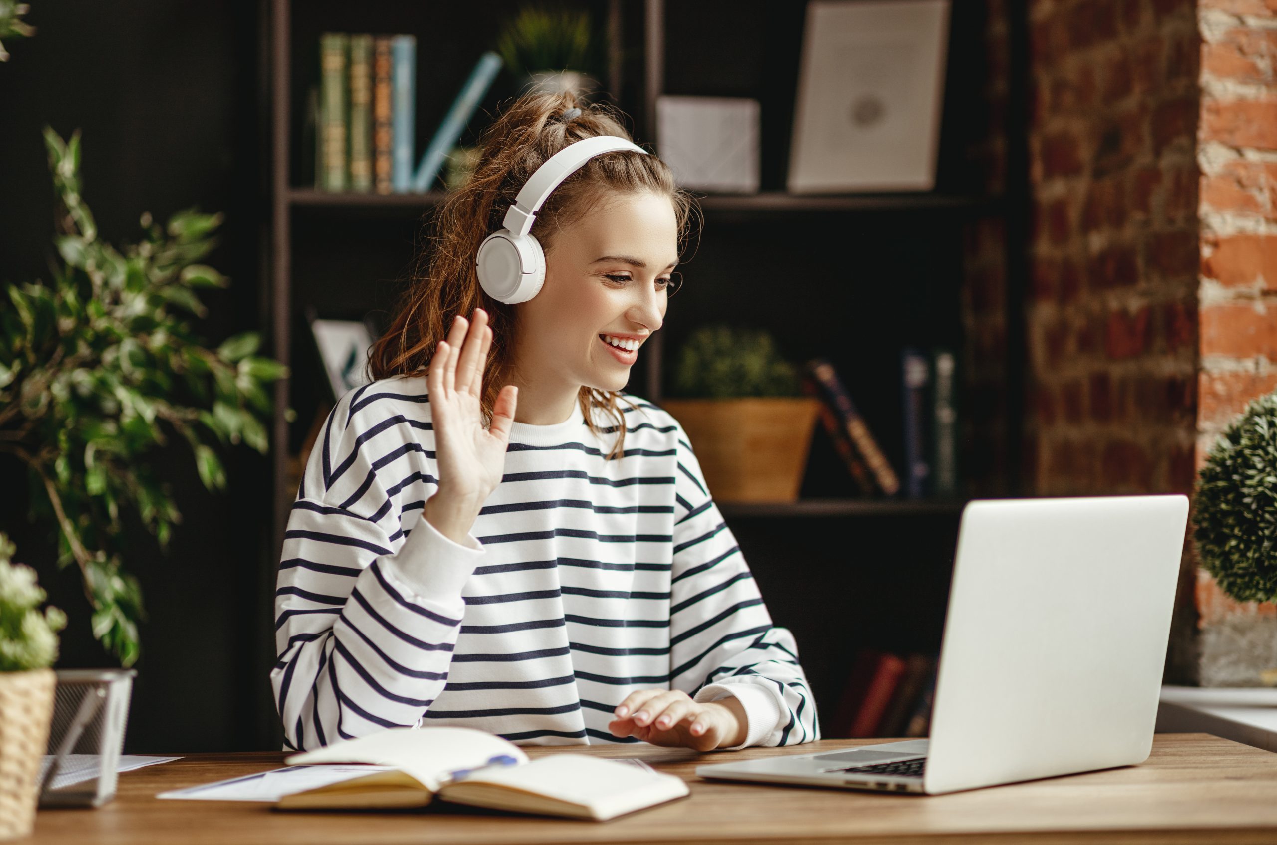 Joyful young female in wireless headphones waving wand greeting to screen while sitting at table and having video chat with business partners using laptop against blurred dark interior of comfortable loft office